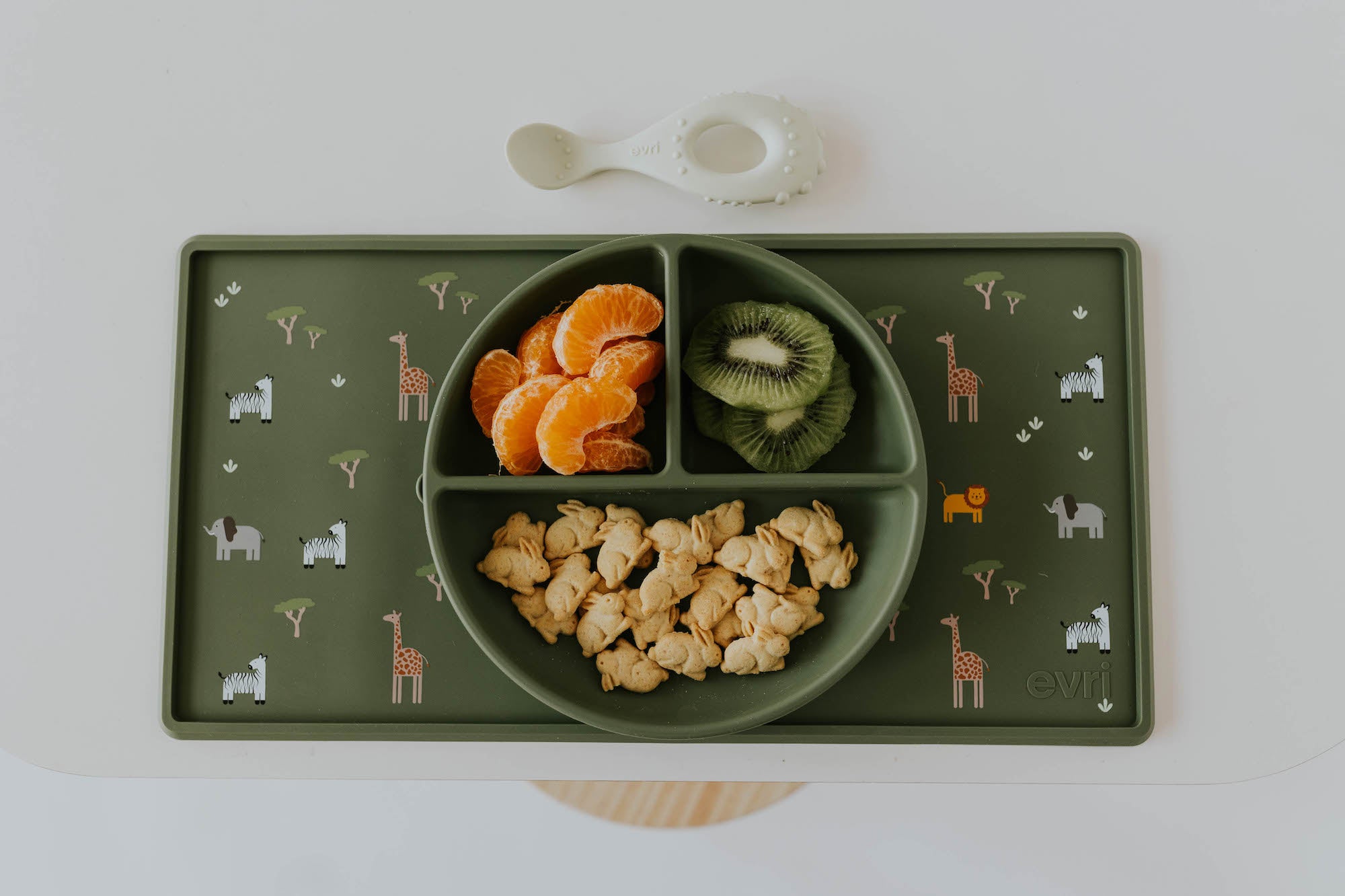 Green silicone divider plate filled with oranges, kiwi, and bunny shaped crackers, sitting on top of a green silicone placemat that features a safari animal print. There is an ivory baby spoon placed just above the placemat. All products designed and sold by Evri.